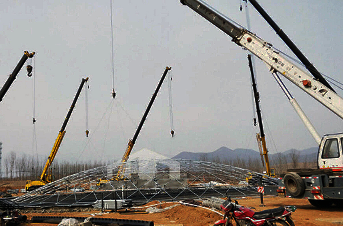 Geodesic dome installation requiring four cranes to lift steel frame structure, showing heavy equipment and complex assembly required for large rigid dome construction