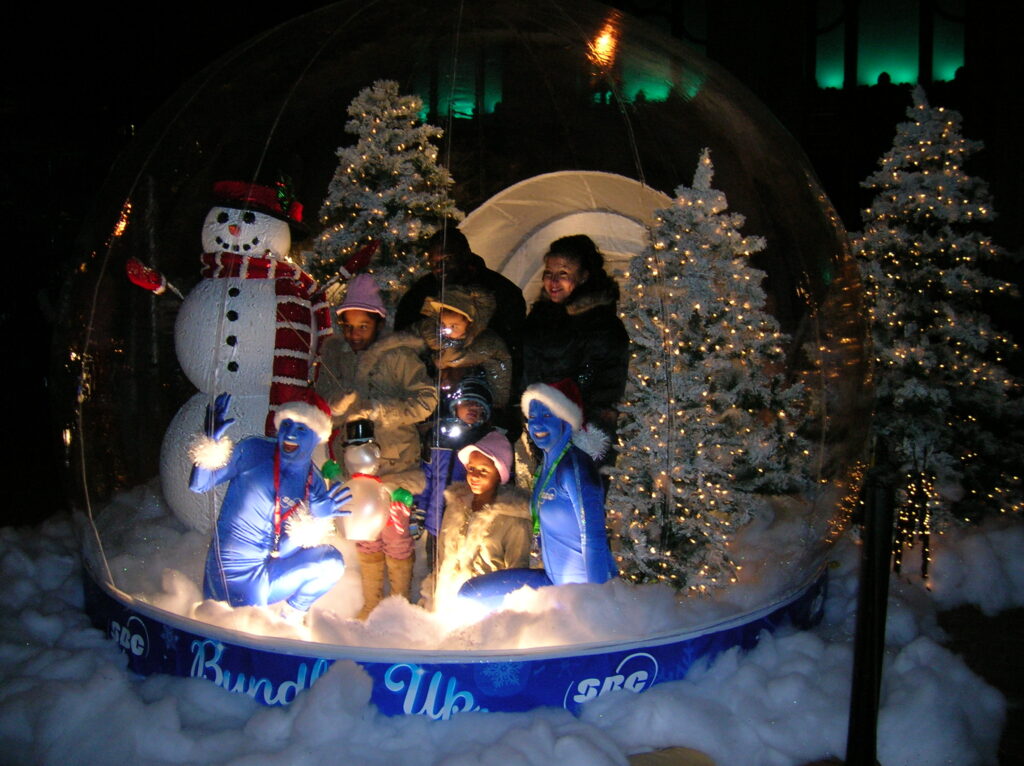 Human snow globe at the SBC Bundle Up holiday activation — a family with young children inside on the rigid flat floor, performers at the front