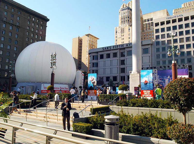 Projection dome exterior daytime — white inflatable projection dome deployed in San Francisco Union Square with Saks Fifth Avenue and Tiffany and Co visible, Hong Kong tourism campaign, pedestrians showing scale