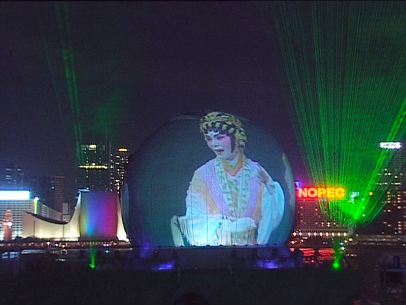 Projection dome exterior night — full 360 degree video projection of Chinese opera performer on inflatable dome surface, deployed on barge in Hong Kong harbor with city skyline and laser display