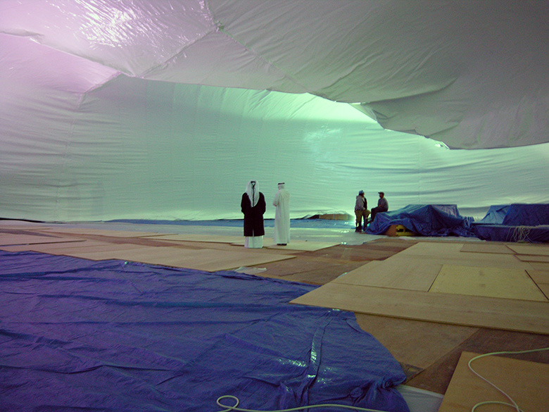 Projection dome interior during early inflation stage — two Gulf clients in traditional dress inspecting dome structure as it rises around them, Doha Qatar