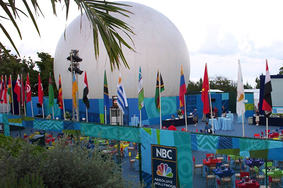 White projection dome at an NBC Olympics broadcast venue, ringed by the international flags of competing nations in daylight