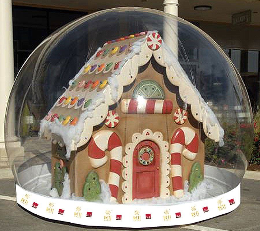 Full-size decorated gingerbread house standing inside a 12 ft snow globe rental outside a shopping center colonnade