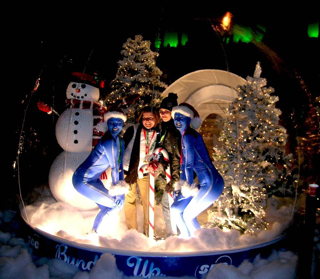 Blue-lit winter scene with a snowman, flocked trees and guests inside a Christmas snow globe rental at a holiday event