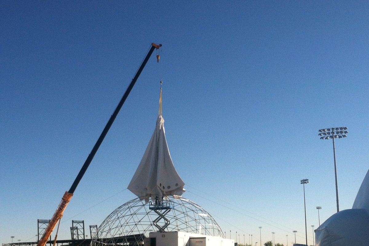 Bare aluminum geodesic dome frame with crane and scissor lift hoisting the cover into place, inflated air dome alongside