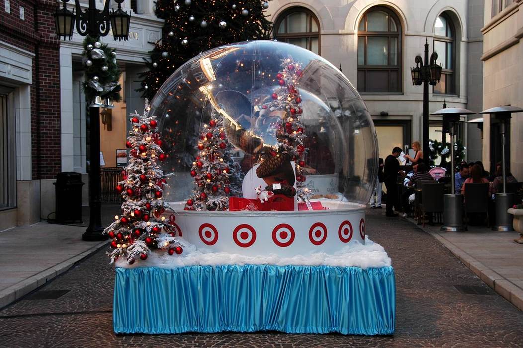 Giant snow globe rental with a sponsor-branded base and lit flocked trees on Rodeo Drive at dusk, diners behind