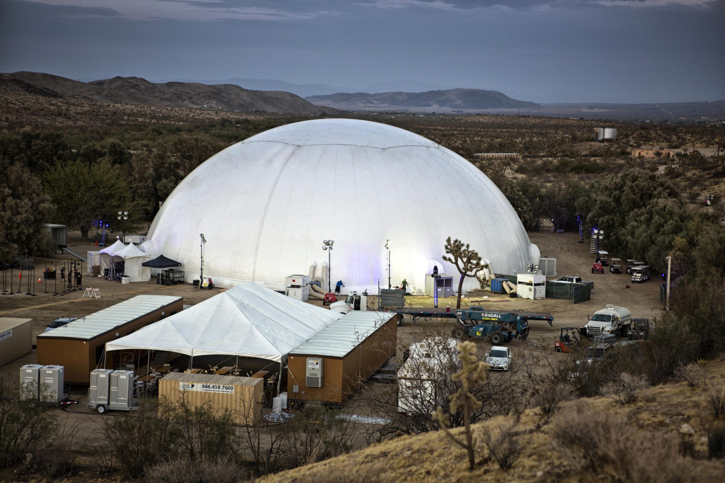Aerial view of a 160 ft projection dome rental installed in open desert near Joshua Tree at golden hour, with production trailers and crane on site.