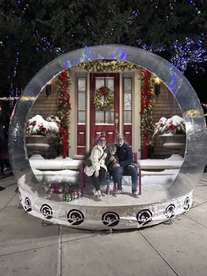 Printed backdrop installed and lit inside a giant snow globe at night, showing the red door holiday scene with a bench in front